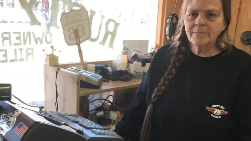 Shop owner in a black T-shirt with her long, brown hair in a single braid cascading down the front of her shirt. She is standing before an old electric cash register with with a storefront window behind her.