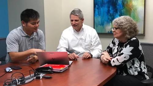 Three office employees gathered around a single laptop in a meeting room.