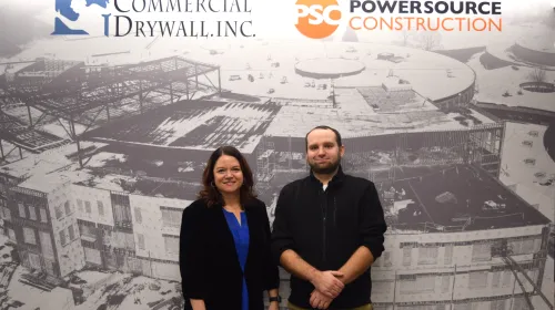Heather Hoffman and Andrew Leach stand in front of a Commercial Drywall, Inc banner, smiling.