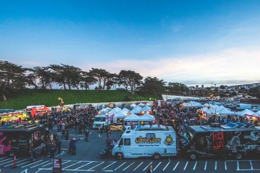 People milling about food trucks and tents in a large parking lot near the ocean