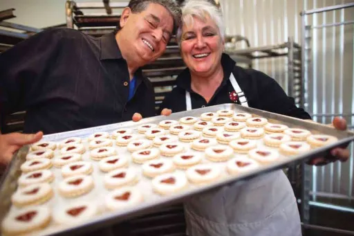 At their new bakery, Fernanda and Angelo Capraro hold a sheet pan of cookies with a red fruit center.