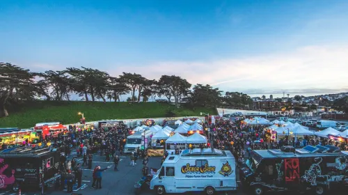 People milling about food trucks and tents in a large parking lot near the ocean