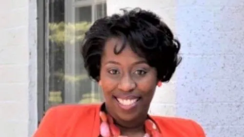 A photo of Donisha Nesbitt smiling at the camera, wearing an orange blazer and matching orange necklace and earrings.