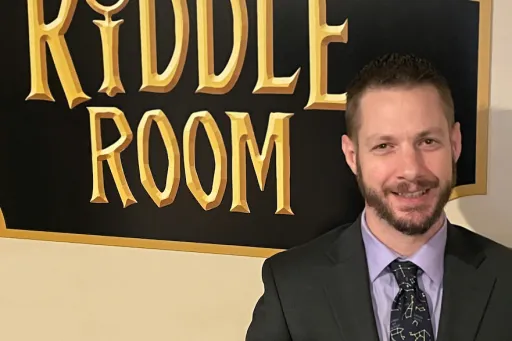 Byron Delmonico standing in front of Riddle Room sign